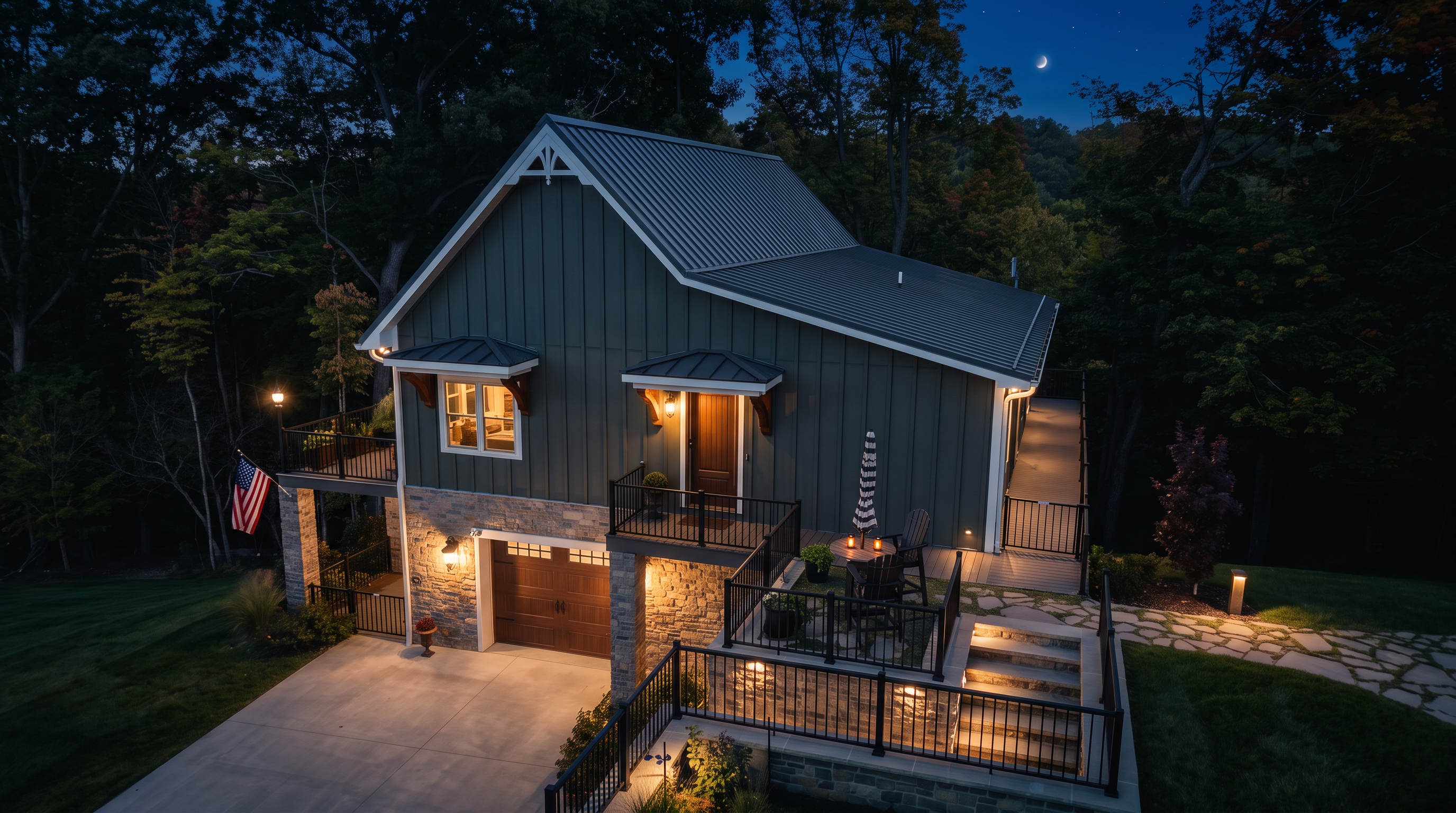 This charming modern farmhouse-style home is captured at twilight, showcasing its striking combination of olive green vertical siding and stone masonry. The multi-level structure features a well-lit garage, an elevated entry porch, and an inviting stone patio area, perfectly accented by warm exterior lighting. Nestled among mature trees with a crescent moon overhead, the home presents a welcoming and picturesque curb appeal.