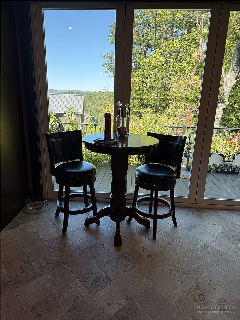 This cozy breakfast nook features a small, round wooden pedestal table paired with two dark leather swivel bar stools. The area is framed by large glass doors that provide an expansive view of the lush, tree-filled landscape and distant mountains beyond. The space is finished with neutral-toned, textured tile flooring, creating a serene and inviting spot for morning coffee.