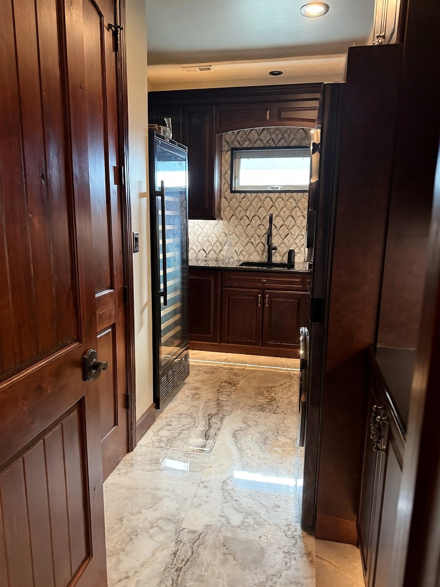This view captures a sophisticated butler's pantry or wet bar area featuring dark wood cabinetry and a tall wine refrigerator. The space is accented by a patterned tile backsplash, a dark sink fixture, and polished stone flooring with intricate veining. The perspective looks through a doorway into the compact, elegant service area, highlighting a blend of luxury finishes and functional design.