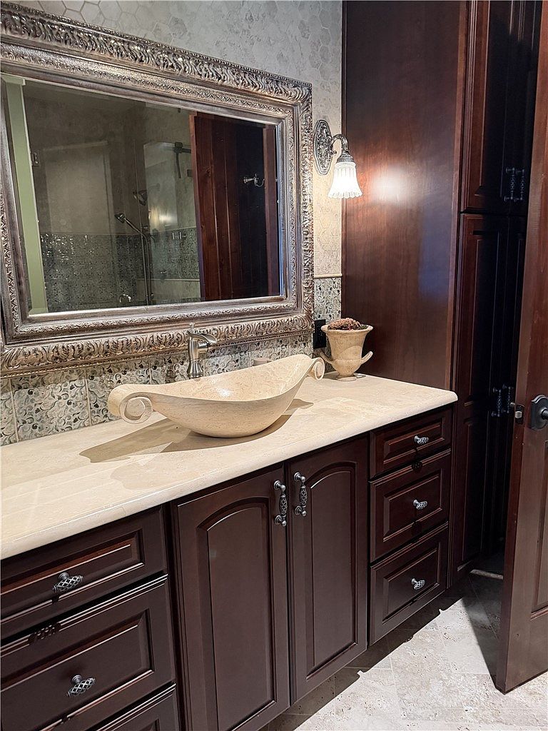 This bathroom features a unique, boat-shaped vessel sink set atop a light-toned stone countertop. The vanity is crafted from dark wood cabinetry with ornate hardware, complemented by a large, decorative framed mirror and a wall-mounted light fixture. The space is finished with patterned tile backsplashes and neutral wall textures, creating a sophisticated and traditional aesthetic.
