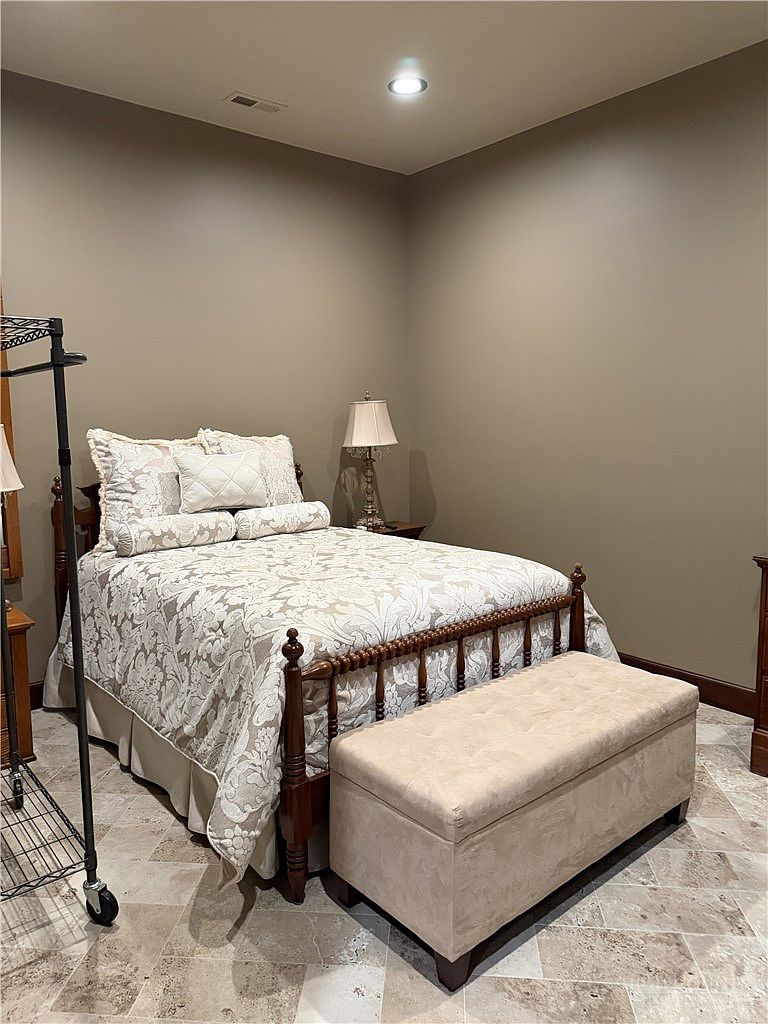 This bedroom features a traditional wooden bed frame with a patterned comforter and a matching upholstered storage bench at the foot. The room is finished with neutral-toned walls and light-colored stone tile flooring, creating a calm and cohesive aesthetic. A bedside lamp provides warm lighting, while a metal garment rack stands to the left, suggesting a functional and well-utilized space.