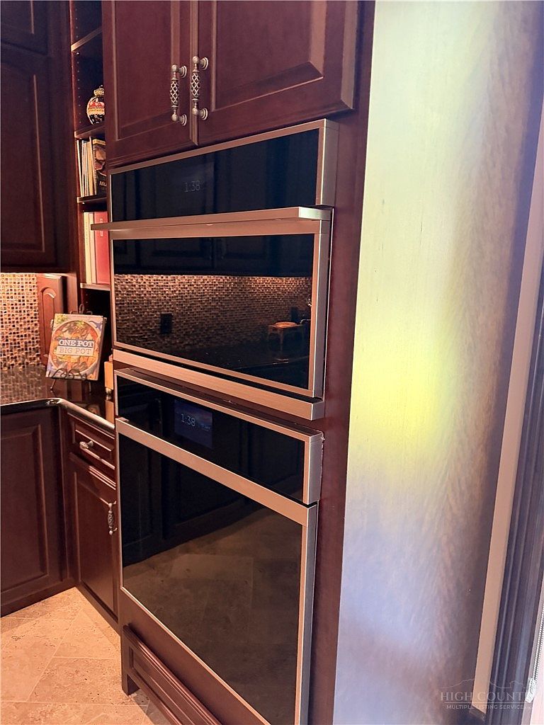 This image captures a built-in wall oven and microwave unit set within dark wood cabinetry in a kitchen. The stainless steel appliances feature sleek, dark glass fronts and digital displays, creating a modern and cohesive look. To the left, a small open shelving area displays a cookbook, adding a touch of personality to the functional space.