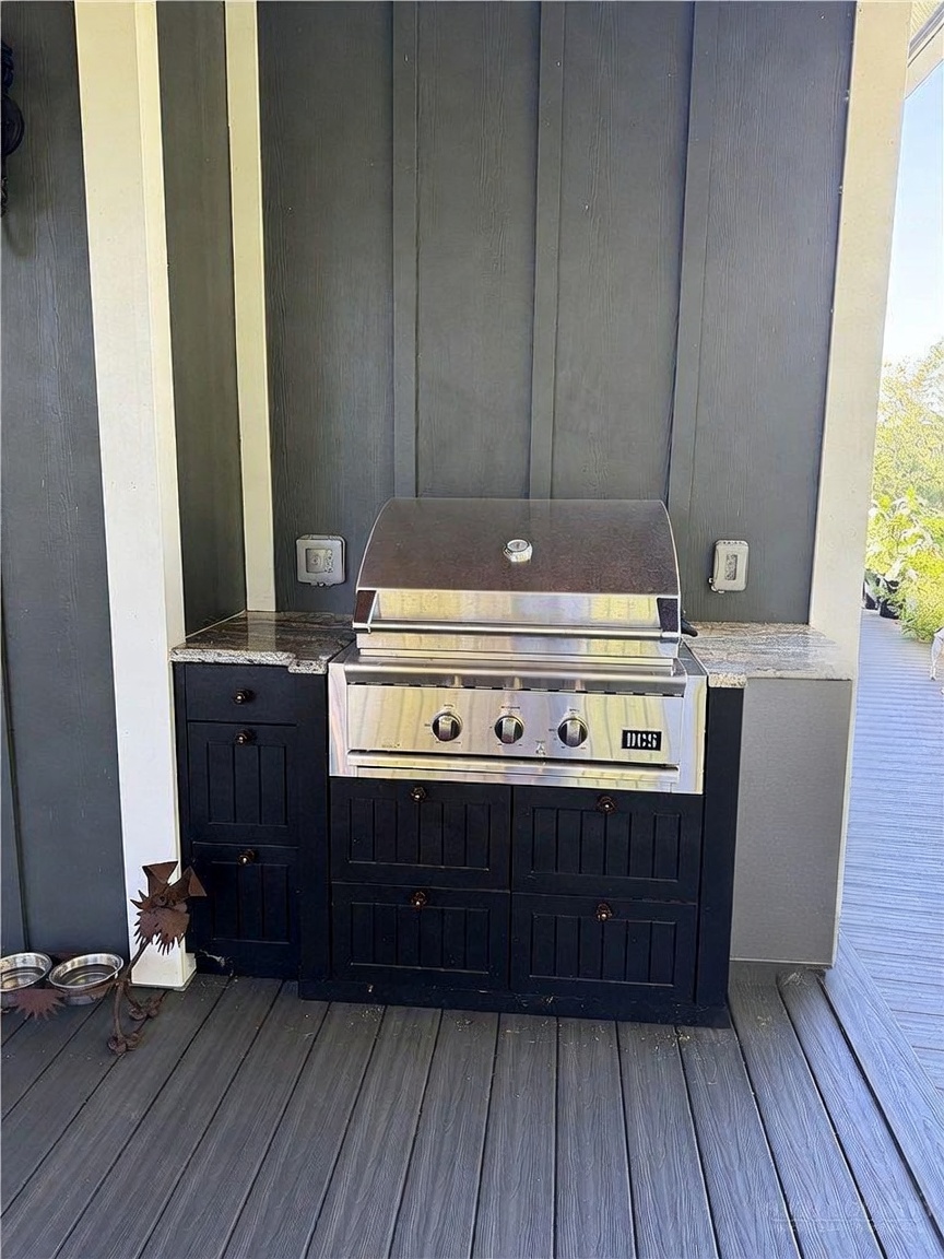 This outdoor kitchen area is situated on a wooden deck, featuring a built-in stainless steel grill set into a dark-painted cabinet base with stone countertops. The setup is framed by vertical dark-toned siding and includes two electrical outlets mounted on the wall. A small, decorative metal sculpture and pet bowls sit on the deck floor to the left, creating a functional and inviting outdoor cooking space.