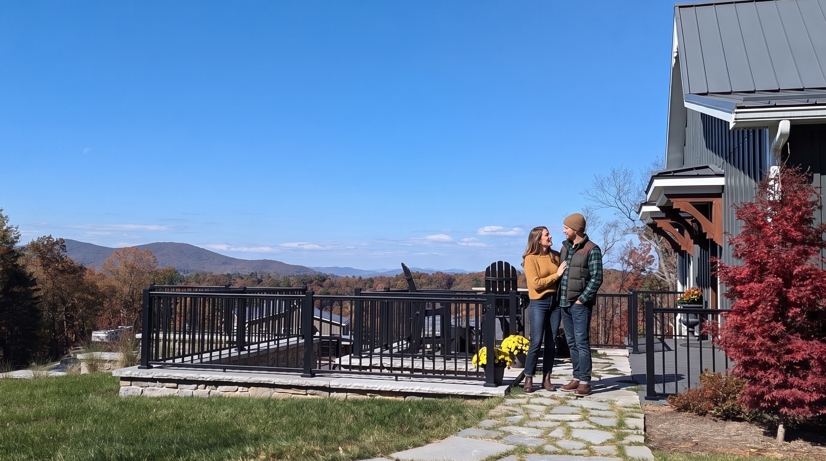 This image captures a spacious outdoor deck featuring a black metal railing and a stone-paved patio area, offering expansive, panoramic views of rolling mountains and autumn-colored trees. A stone walkway leads toward the deck, which is furnished with dark chairs and accented by potted yellow flowers. The scene is set against a clear blue sky, highlighting the home's modern siding and architectural wood detailing on the right side.