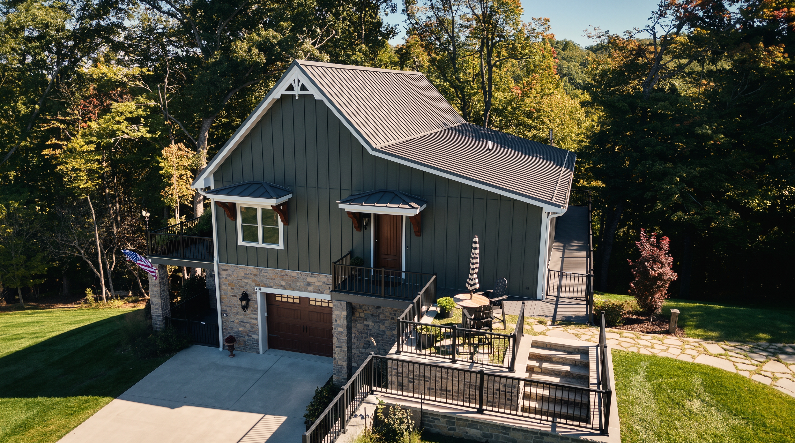 This elevated perspective captures a modern, two-story home with a distinctive dark vertical siding and a metal roof. The lower level features stone masonry, a garage, and an expansive multi-level deck with black railings that overlooks a lush, wooded landscape. A stone pathway leads toward the house, emphasizing a serene, private setting surrounded by mature trees.