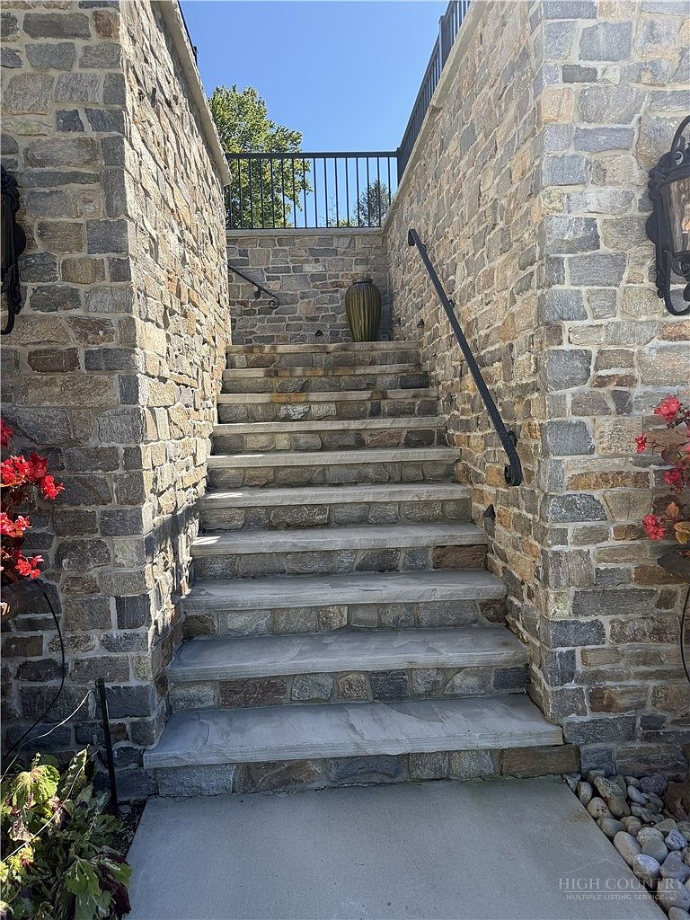 This exterior entryway features a set of stone steps leading upward, flanked by rustic stone walls on both sides. A black metal railing provides safety along the staircase, which is accented by decorative floral arrangements and a tall, textured vase at the landing. The scene is captured from a low, eye-level perspective, emphasizing the solid masonry construction and the inviting, classic aesthetic of the home's approach.