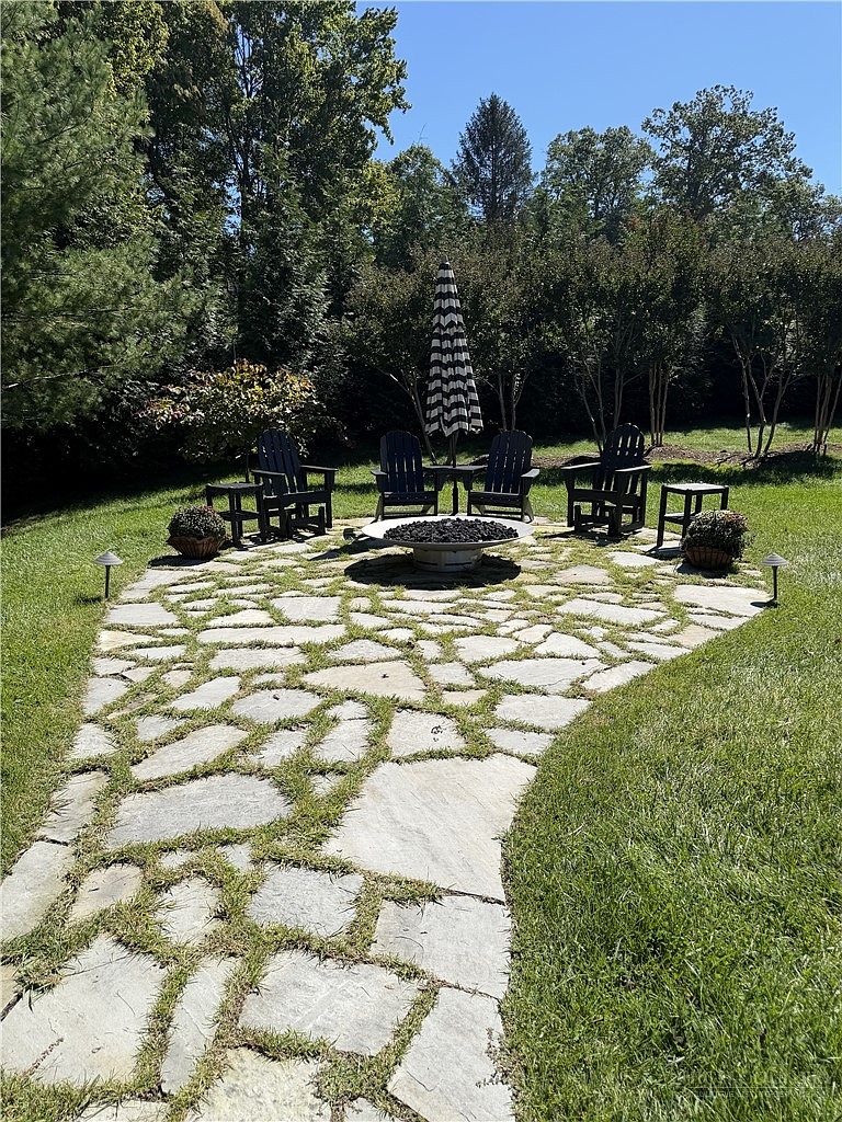 This outdoor living space features a flagstone patio with a central fire pit, surrounded by four dark Adirondack chairs and two side tables. A striped umbrella provides shade, and the area is framed by lush green trees and manicured lawn. The perspective is a ground-level shot looking toward the seating area, creating an inviting and tranquil atmosphere for relaxation.