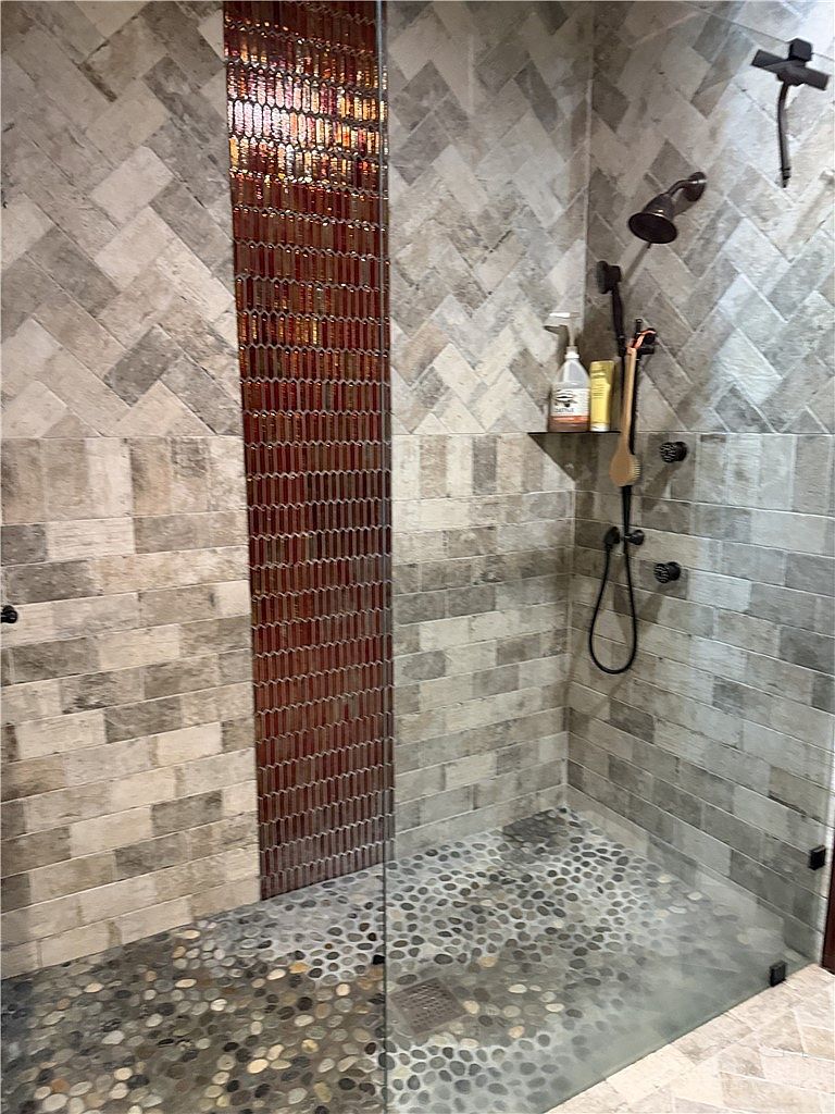 This shower features a distinctive design with a vertical accent strip of iridescent, narrow tiles flanked by sections of herringbone-patterned stone and horizontal rectangular tiles. The shower floor is finished with smooth, rounded river stones, and the space is equipped with dark bronze fixtures, including a rainfall showerhead and a handheld sprayer. A glass panel encloses the shower, providing a clear view of the textured wall treatments and the built-in corner shelf.