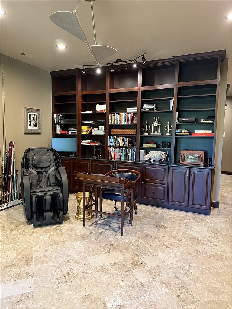 This home office features a large, dark wood built-in bookshelf unit that spans the wall, providing ample storage and display space. A professional-grade massage chair sits to the left, while a classic wooden desk and chair are positioned in the center of the room. The space is finished with neutral-toned stone tile flooring and recessed lighting, creating a sophisticated and functional workspace.