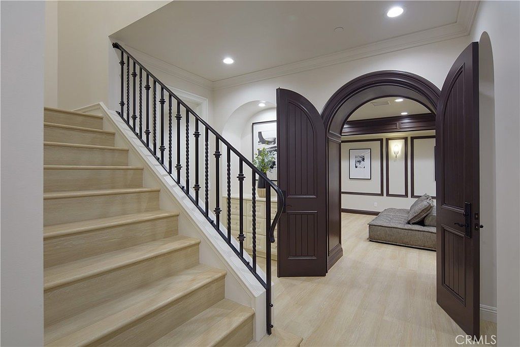 This interior view captures a grand staircase with light wood treads and a dark metal railing leading to an upper level. To the right, a pair of dark, arched double doors opens into a room featuring paneled walls and a low-profile seating area. The space is bright and neutral, showcasing a blend of traditional architectural elements and modern light-toned flooring.