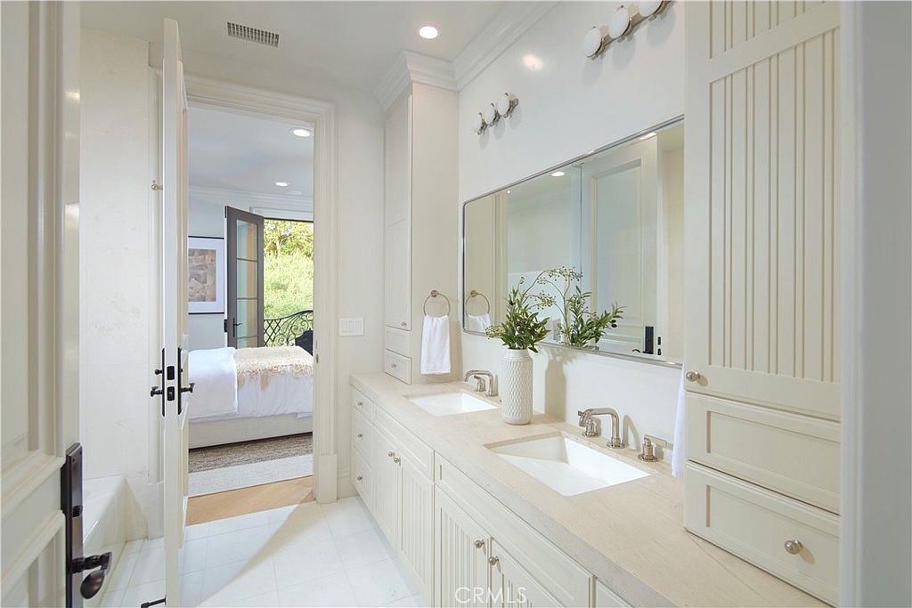 This bright and elegant primary bathroom features a dual-sink vanity with light-toned cabinetry and a long, rectangular mirror. The space is finished with clean, neutral surfaces and includes a doorway leading directly into an adjacent bedroom. The perspective is captured from the doorway, highlighting the seamless transition between the private bath and the sleeping area.