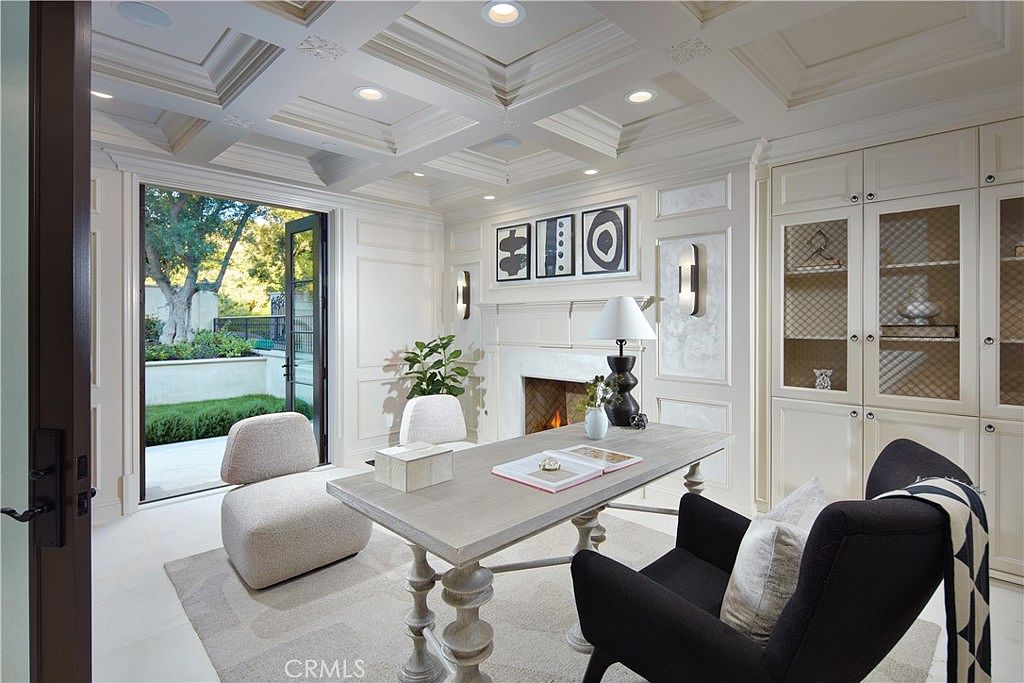 This sophisticated home office features a coffered ceiling, elegant wall paneling, and a light-toned desk centered in the room. A fireplace with a classic mantel serves as a focal point, flanked by built-in cabinetry with glass-front doors. Large glass doors open to an outdoor patio, allowing natural light to fill the space and creating a seamless transition between indoor and outdoor environments.