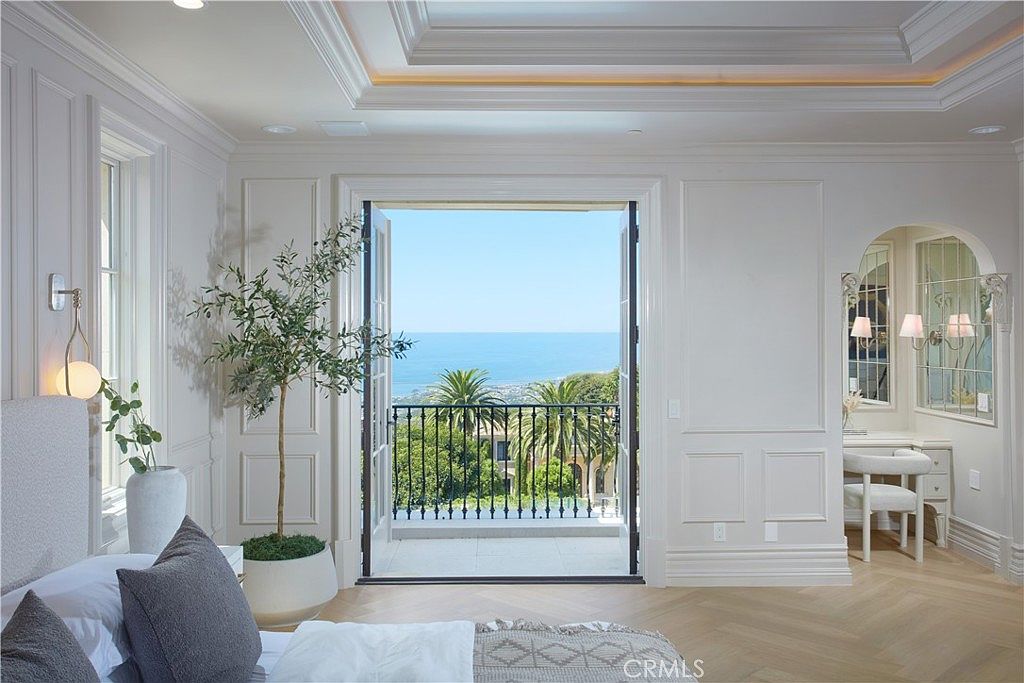 This elegant primary bedroom features light wood herringbone flooring and crisp white walls with classic wainscoting. A set of French doors opens onto a private balcony, framing a stunning, unobstructed view of the ocean and lush palm trees. To the right, a built-in vanity area with a mirror and soft lighting adds a touch of luxury to the bright, airy space.