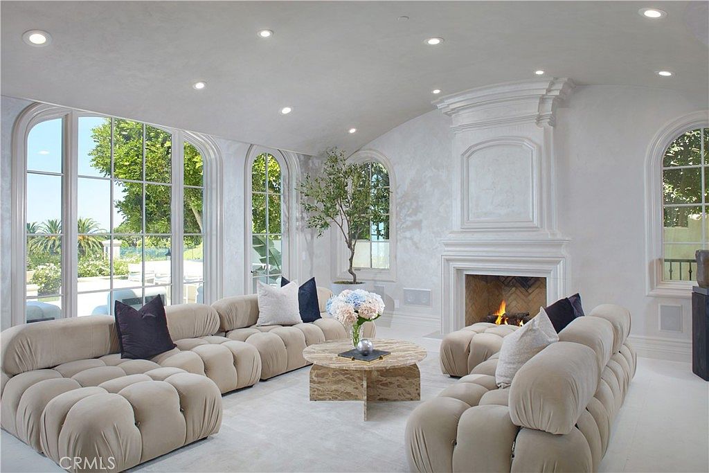 This elegant living room features a soft, neutral color palette with plush, modular seating arranged around a circular stone coffee table. A prominent fireplace with a tall, decorative surround serves as a focal point, while large arched windows allow for abundant natural light and views of the outdoors. The space is accented by a potted tree and recessed lighting, creating a sophisticated and airy atmosphere.