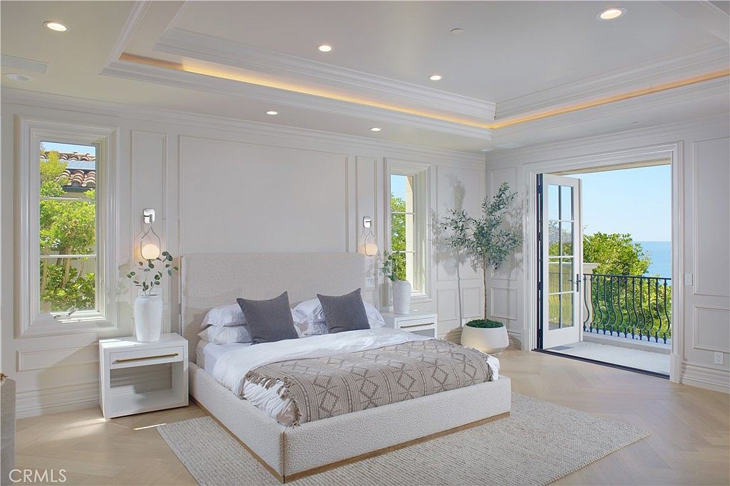 This elegant primary bedroom features a neutral color palette, light wood flooring, and detailed wall molding that adds a sophisticated touch. A plush bed with a textured headboard sits centered between two matching nightstands, while a pair of glass doors opens to a private balcony with ocean views. The room is bathed in natural light from the balcony and side windows, creating a serene and airy atmosphere.