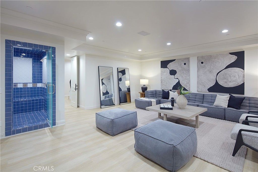 This spacious living area features a large, modular grey sectional sofa and two matching ottomans arranged on a light-toned area rug. The room is accented by two tall, leaning floor mirrors and two large, abstract black-and-white art pieces mounted on the wall. To the left, a unique glass-enclosed shower stall with vibrant blue tiles adds a bold, unexpected design element to the open-concept space.