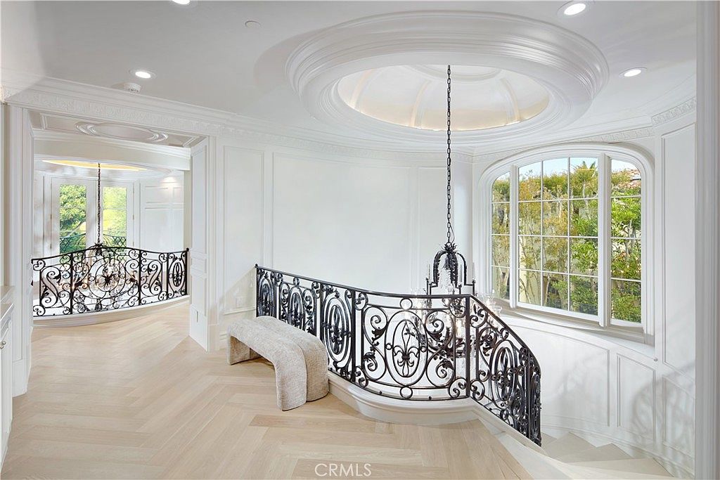This elegant landing area features light wood flooring laid in a herringbone pattern and a dramatic curved staircase with intricate black wrought-iron railings. A large, circular recessed ceiling detail with a chandelier serves as a focal point, while tall, arched windows flood the space with natural light. The neutral white walls and classic architectural molding create a sophisticated and airy atmosphere.