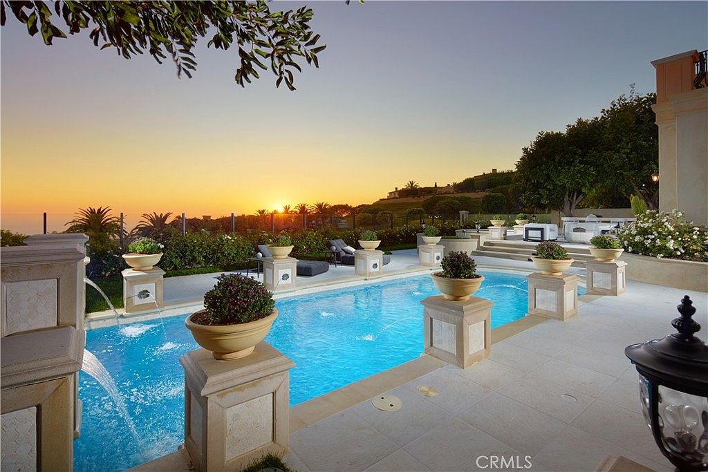 This luxurious outdoor space features a rectangular swimming pool lined with decorative stone pedestals topped by potted plants. The pool area is paved with light-colored stone tiles and overlooks a scenic hillside landscape during a vibrant sunset. The setting is elegant and serene, emphasizing a high-end lifestyle with a clear view of the horizon.