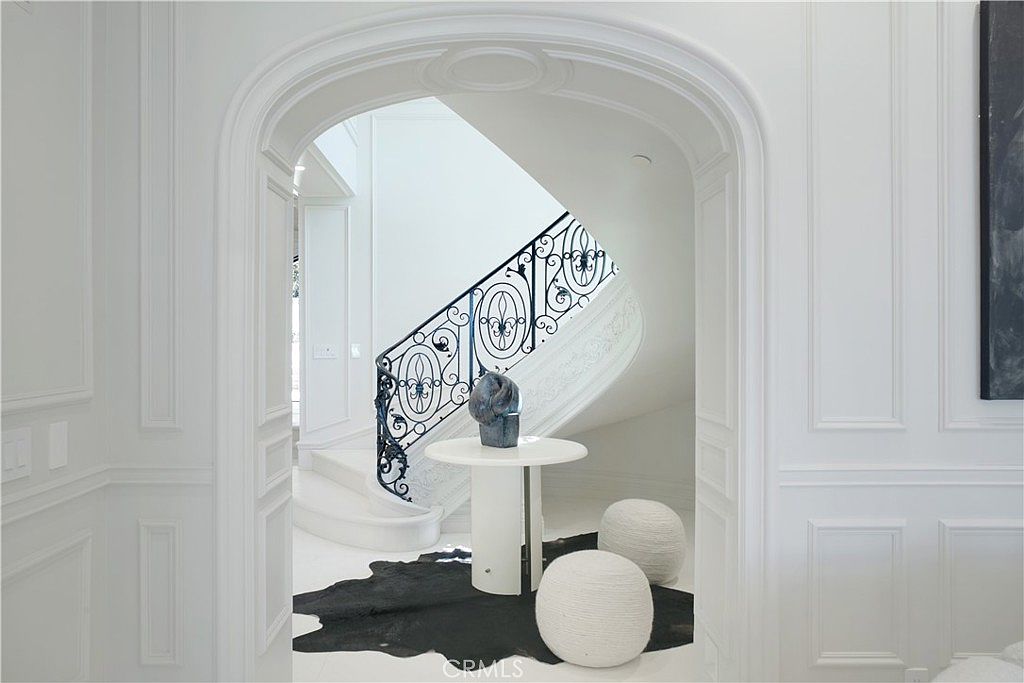 This elegant entryway features a grand, curved staircase with intricate black wrought-iron railings, framed by a classic arched doorway. A minimalist white pedestal table sits atop a dark, organic-shaped rug, accented by two textured, spherical ottomans. The space is defined by crisp white walls with detailed wainscoting, creating a sophisticated and airy atmosphere.