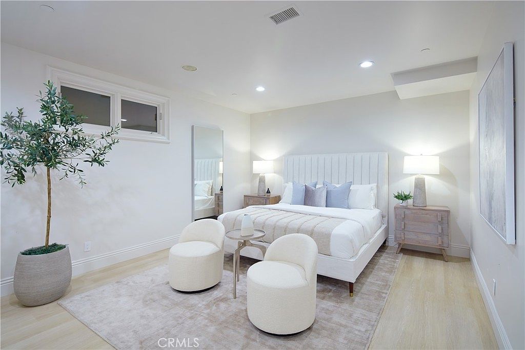 This bright and airy primary bedroom features a neutral color palette with light wood flooring and a large area rug. The room is anchored by a tall, upholstered headboard flanked by two matching nightstands with lamps, while a pair of rounded accent chairs and a small table create a cozy seating area. A large potted olive tree adds a touch of greenery, and a full-length mirror leans against the wall, enhancing the sense of space in this modern, serene retreat.