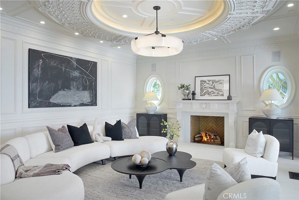 This elegant living room features a sophisticated monochromatic palette with a large, curved white sectional sofa and a unique, multi-tiered black coffee table. The space is anchored by a traditional fireplace with a detailed mantel and a herringbone-patterned firebox, complemented by symmetrical arched windows and ornate ceiling molding. The room exudes a refined, contemporary aesthetic, enhanced by abstract artwork and a textured area rug.