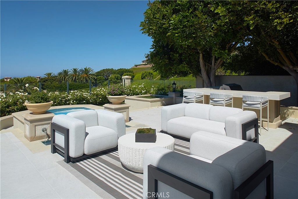 This expansive outdoor patio features a modern seating arrangement with white upholstered furniture set atop a striped area rug. The space includes a built-in bar area, a small spa, and lush landscaping, all set against a backdrop of rolling hills and a clear blue sky. The perspective is a wide-angle shot from the patio, emphasizing the seamless blend of luxury outdoor living and scenic views.