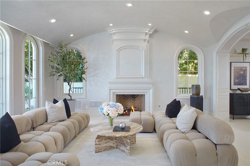 This elegant living room features a symmetrical layout centered around a fireplace with a prominent, tall white mantel surround. The space is furnished with two large, tufted modular sofas in a neutral tone facing a circular stone coffee table, creating a sophisticated and comfortable seating area. Arched windows on either side of the fireplace provide natural light, while a potted tree adds a touch of greenery to the bright, airy atmosphere.