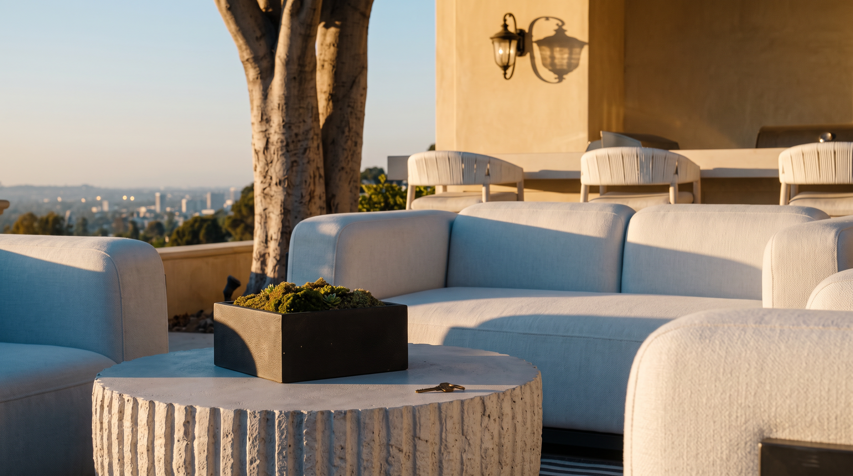 This outdoor patio space features a modern, light-toned seating arrangement centered around a textured, cylindrical coffee table on a striped area rug. In the background, a built-in bar area with three stools sits beneath the shade of mature, lush trees, creating a serene and private atmosphere. The scene is captured from a low, eye-level perspective, emphasizing the comfortable lounge area and the seamless transition between the indoor living space and the landscaped backyard.