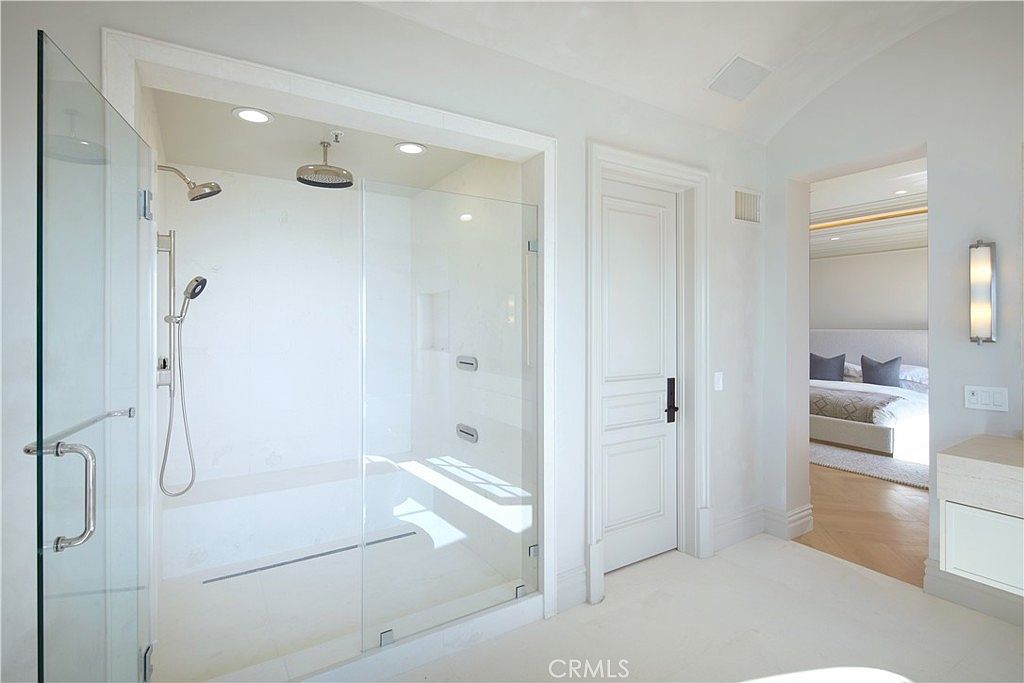 This bright, modern bathroom features a spacious walk-in shower enclosed by glass panels, complete with both a rainfall showerhead and a handheld sprayer. A white door leads to an adjacent bedroom, which is visible through the doorway, showcasing a neutral color palette and clean lines. The space is finished with light-toned stone surfaces and minimalist fixtures, creating a serene and luxurious atmosphere.