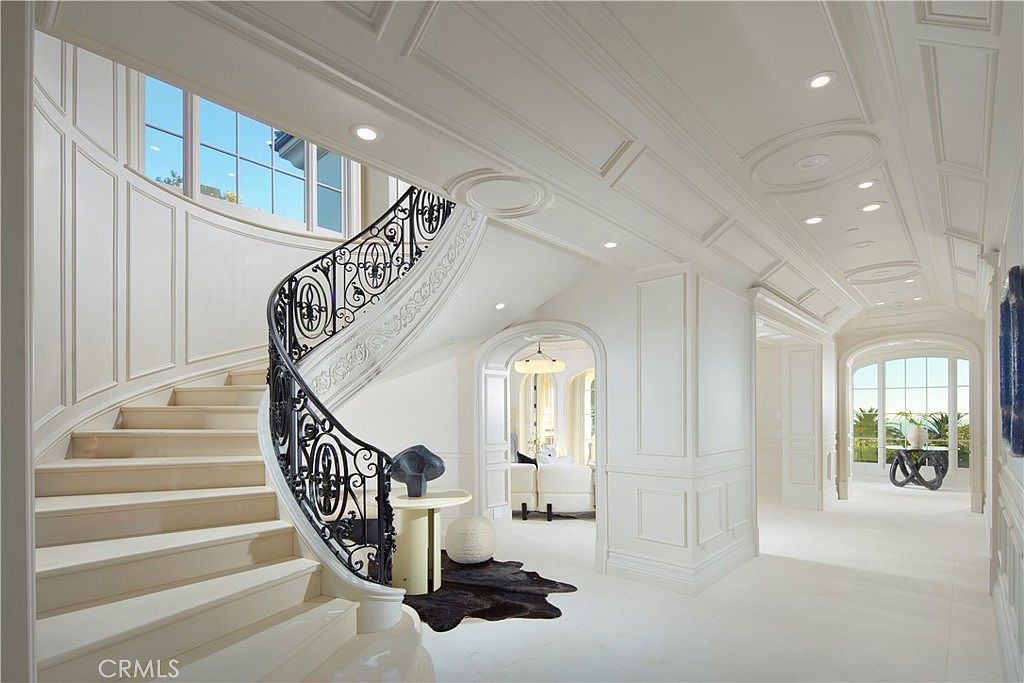 This grand foyer features a sweeping curved staircase with an intricate black metal railing and light-toned stone steps. The space is characterized by elegant wall paneling, a coffered ceiling with recessed lighting, and a bright, airy atmosphere. A small pedestal table with a sculptural piece sits at the base of the stairs, leading the eye toward an arched doorway that opens into a sunlit living area.