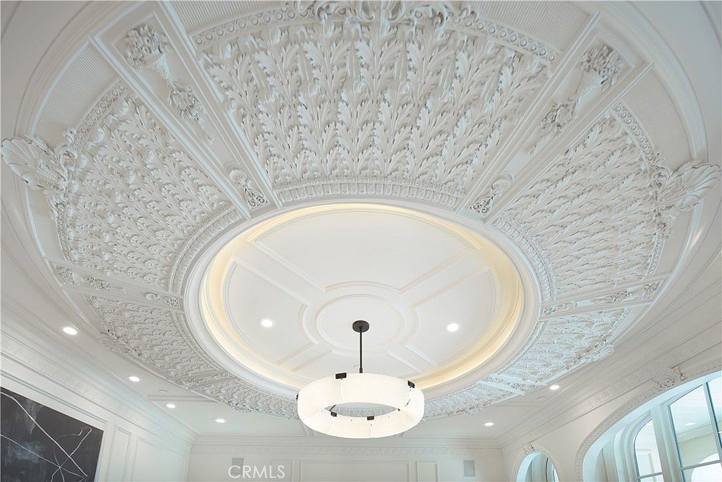 This image captures an elaborate, circular ceiling medallion featuring intricate, carved leaf-like molding that radiates outward from a central recessed dome. A modern, cylindrical chandelier with a textured fabric shade hangs from the center, providing a contemporary contrast to the ornate, classical plasterwork. The perspective is looking directly upward, emphasizing the grandeur and architectural craftsmanship of the ceiling design.