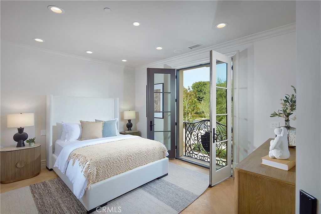 This bright and airy bedroom features a neutral color palette with a plush white bed, light wood flooring, and a textured area rug. The room is highlighted by a set of French doors that open onto a private balcony, allowing for ample natural light and a view of the surrounding greenery. The space is accented by minimalist bedside tables, soft lighting, and a decorative sculpture on a wooden dresser, creating a serene and inviting atmosphere.