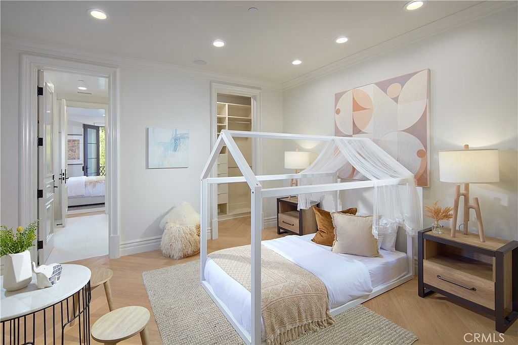 This bright and airy bedroom features a whimsical house-frame bed draped with sheer fabric, set against a neutral color palette with light wood flooring. The room includes a small seating area with a round table, a bedside table with a lamp, and an open-concept walk-in closet visible in the background. A large abstract art piece hangs above the bed, contributing to the room's modern and playful aesthetic.