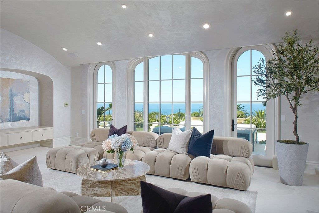 This elegant living room features a plush, modular beige sofa arranged in a U-shape, centered around a round stone coffee table. Large, arched windows provide an expansive view of the ocean and sky, filling the space with natural light. The room is decorated in a soft, neutral palette with textured wall finishes and a potted olive tree, creating a serene and sophisticated atmosphere.