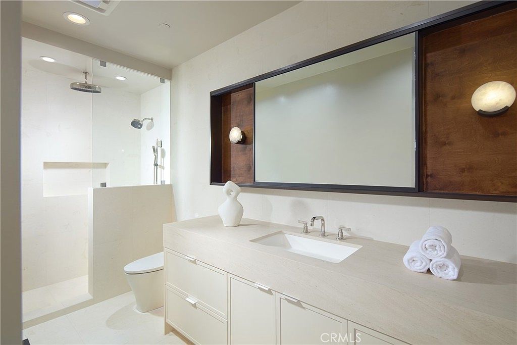 This modern bathroom features a sleek, light-toned vanity with a single integrated sink and a large, rectangular mirror framed in dark wood. To the left, a glass-enclosed walk-in shower with a rainfall showerhead is visible, while a stack of white towels rests on the countertop to the right. The space is illuminated by warm wall-mounted light fixtures, creating a clean and sophisticated atmosphere.