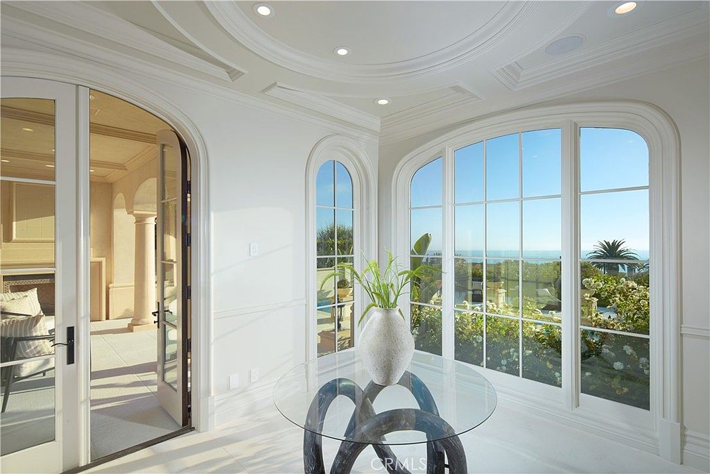 This bright and airy sunroom features a circular glass-topped table as a central focal point, set against a backdrop of expansive, arched windows that offer panoramic ocean views. The room is characterized by its elegant, neutral color palette, curved architectural lines, and a coffered ceiling that adds a sense of sophistication. A set of French doors leads to an adjacent space, enhancing the open and flowing layout of this luxurious home.