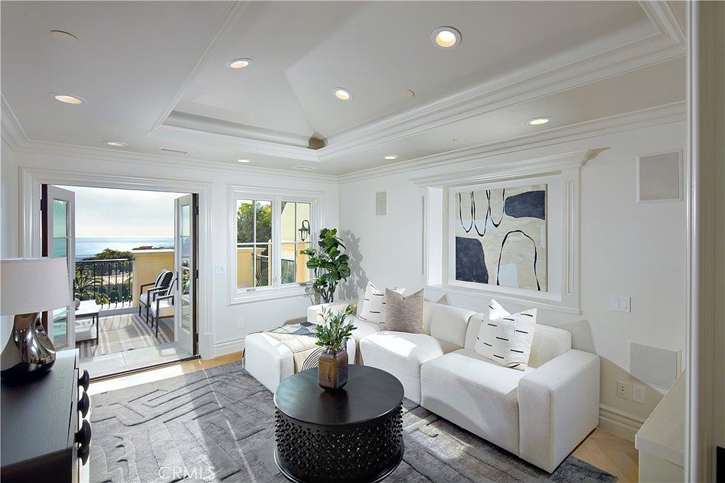 This elegant living room features a bright, neutral color palette with a plush white sectional sofa and a dark, textured circular coffee table. The space is highlighted by a tray ceiling with recessed lighting and large glass doors that open onto a balcony with ocean views. A piece of abstract wall art serves as a focal point above the sofa, creating a sophisticated and airy atmosphere.