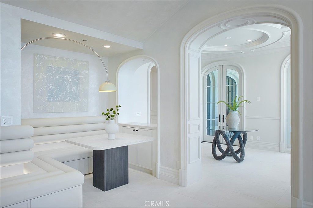 This elegant dining area features a built-in banquette with plush, channel-tufted seating and a modern, minimalist table. An arched doorway leads into a bright, circular foyer, where a glass-topped table with a sculptural base serves as a focal point. The space is defined by a monochromatic white palette, sophisticated architectural arches, and a sleek, curved floor lamp that adds a contemporary touch.