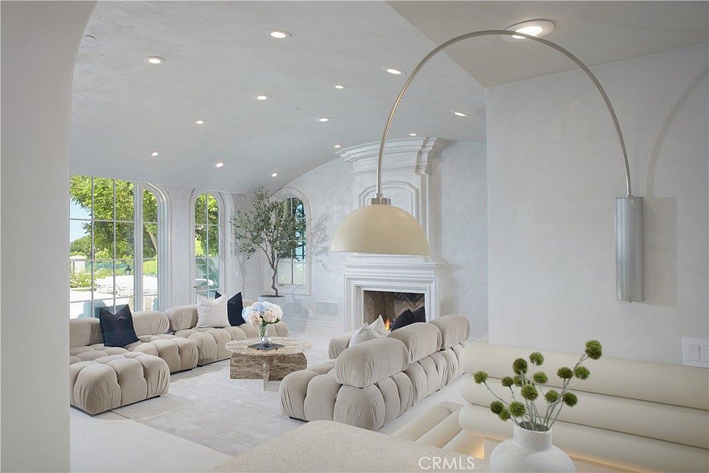 This bright and airy living room features a curved ceiling with recessed lighting and a neutral color palette. The space is anchored by a central fireplace with a decorative mantel and is furnished with plush, modular-style sofas and a round stone coffee table. Large arched windows allow for ample natural light, while a prominent arched floor lamp adds a modern sculptural element to the room.