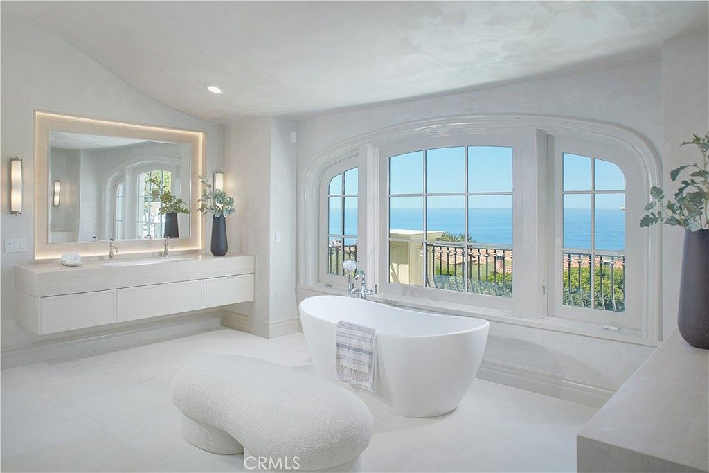 This luxurious primary bathroom features a minimalist aesthetic with a sleek, wall-mounted vanity and a freestanding soaking tub positioned in front of large, arched windows that frame an expansive ocean view. The room is finished in a monochromatic light palette, complemented by a textured, curved bench in the foreground and elegant wall sconces flanking the mirror. The perspective is centered, emphasizing the serene atmosphere and the seamless connection between the indoor space and the coastal scenery.