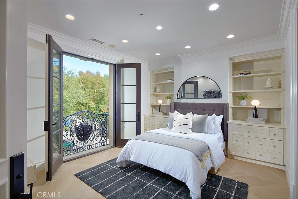 This bright and airy bedroom features light wood flooring and a neutral color palette, centered around a plush bed with a dark upholstered headboard. Built-in shelving units flank the bed, providing ample storage and display space, while a set of French doors opens onto a private balcony with decorative iron railings. The room is illuminated by recessed lighting, creating a clean and inviting atmosphere.