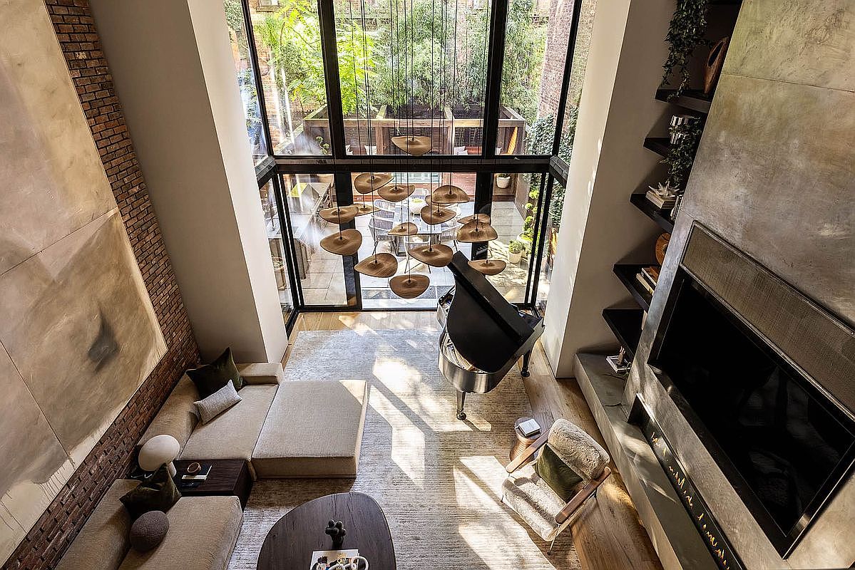 This is a high-angle shot of a luxurious living room featuring a grand piano, a sectional sofa, and a modern fireplace with built-in shelving. The room is bathed in natural light from a large window overlooking a garden, and a unique chandelier adds a touch of elegance. The overall impression is one of sophistication and comfort.