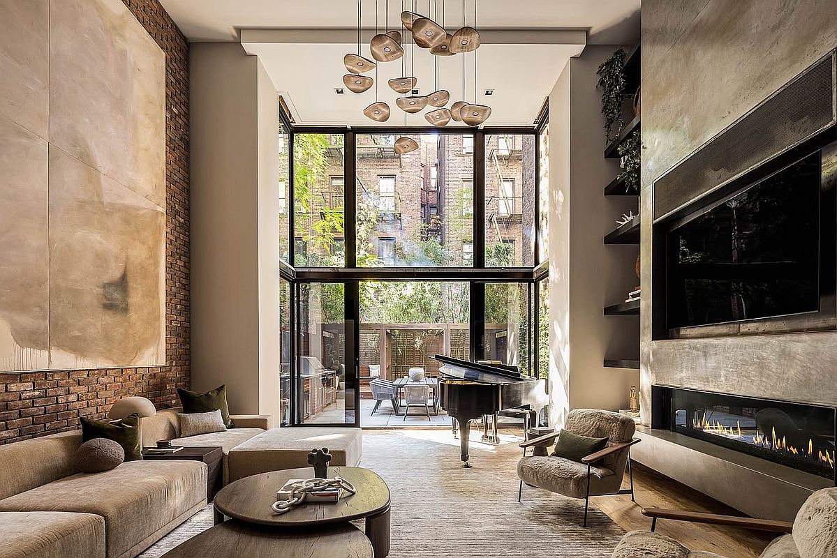 This is a luxurious living room featuring a double-height window wall that provides ample natural light and a view of the outdoor patio and surrounding buildings. The room is furnished with a large sectional sofa, a grand piano, and a modern fireplace with a television above. The style is sophisticated and contemporary, with a focus on natural materials and textures.