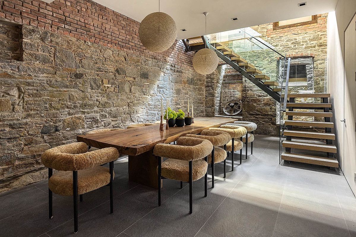 This interior shot showcases a dining room with a rustic yet modern aesthetic. A large, live-edge wooden dining table is surrounded by unique, upholstered chairs with black legs. The room features exposed brick and stone walls, complemented by modern pendant lighting and a staircase with glass railings, creating a blend of old-world charm and contemporary design.