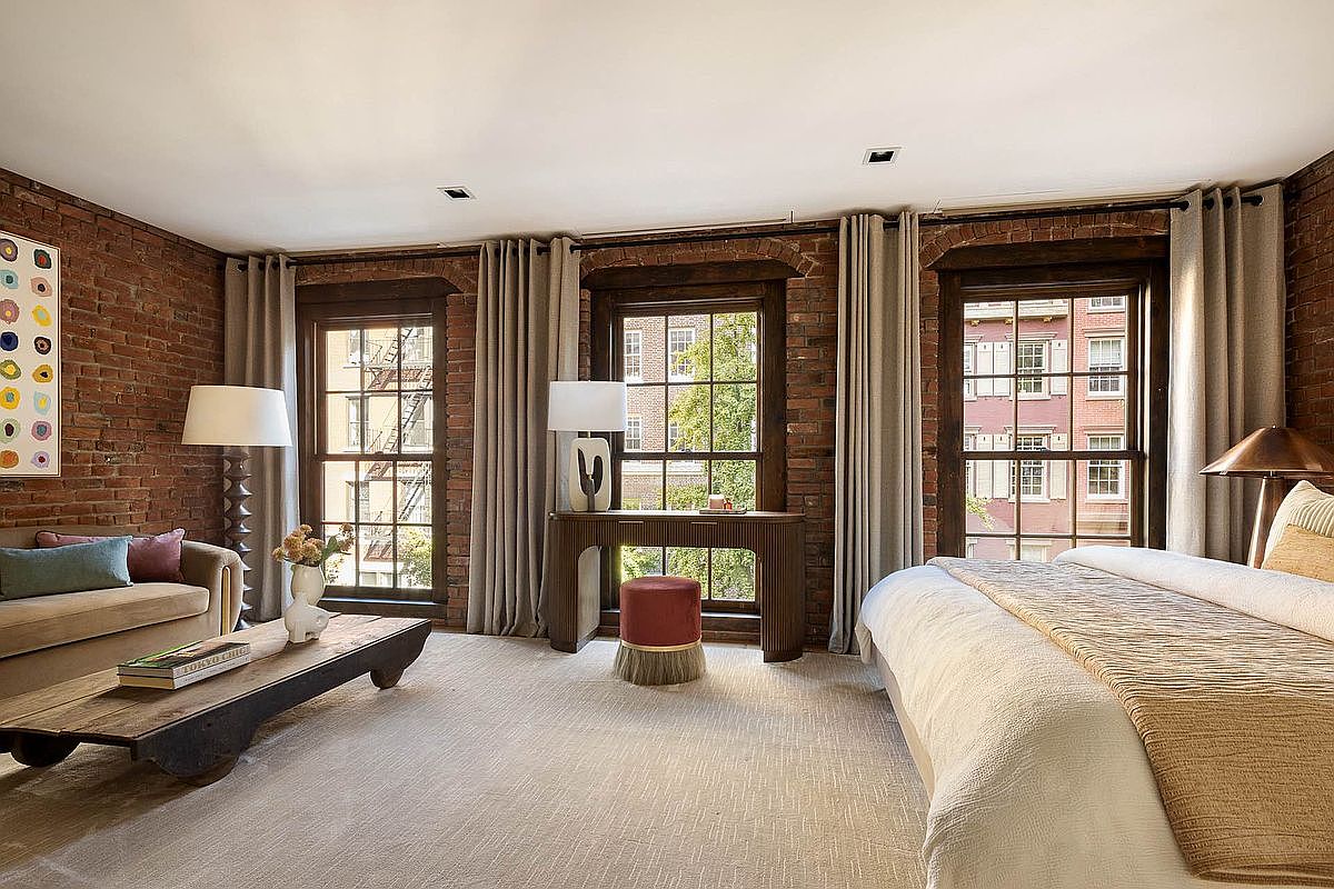 This primary bedroom features exposed brick walls, three large windows with neutral-toned curtains, and a light-colored carpet. The room includes a seating area with a sofa and coffee table, a console table with a lamp, and a king-size bed with a white duvet and tan throw. The overall aesthetic is a blend of rustic and modern, creating a warm and inviting space.