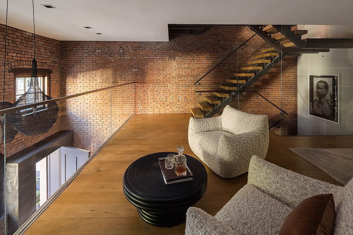 This interior shot showcases a modern living room with exposed brick walls and hardwood floors. A unique staircase leads to another level, while comfortable seating arrangements invite relaxation. A round black coffee table sits in the center, adding a touch of contemporary style to the space.