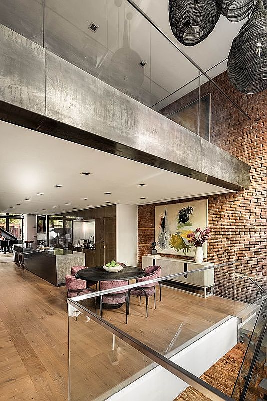 This interior shot showcases a modern dining area with a round table surrounded by upholstered chairs, set on a wooden floor. A large abstract painting hangs on the exposed brick wall, complemented by a sleek sideboard. The open-concept design flows into the kitchen area, creating a spacious and inviting atmosphere.
