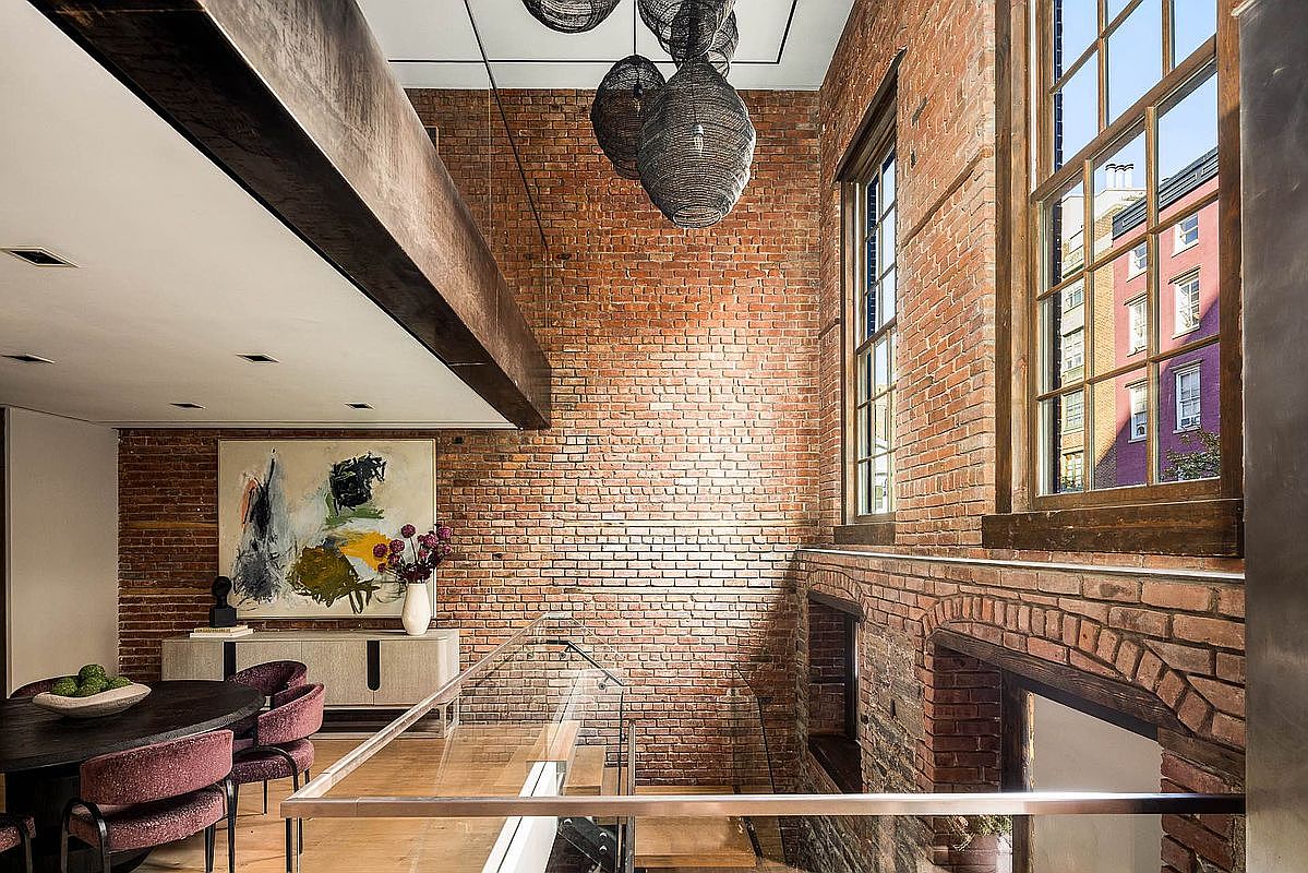 This interior shot showcases a dining area with exposed brick walls, large windows, and modern lighting fixtures. A dark wood dining table is surrounded by upholstered chairs, and an abstract painting hangs above a sideboard. The space exudes a blend of industrial and contemporary design, creating a sophisticated and inviting atmosphere.
