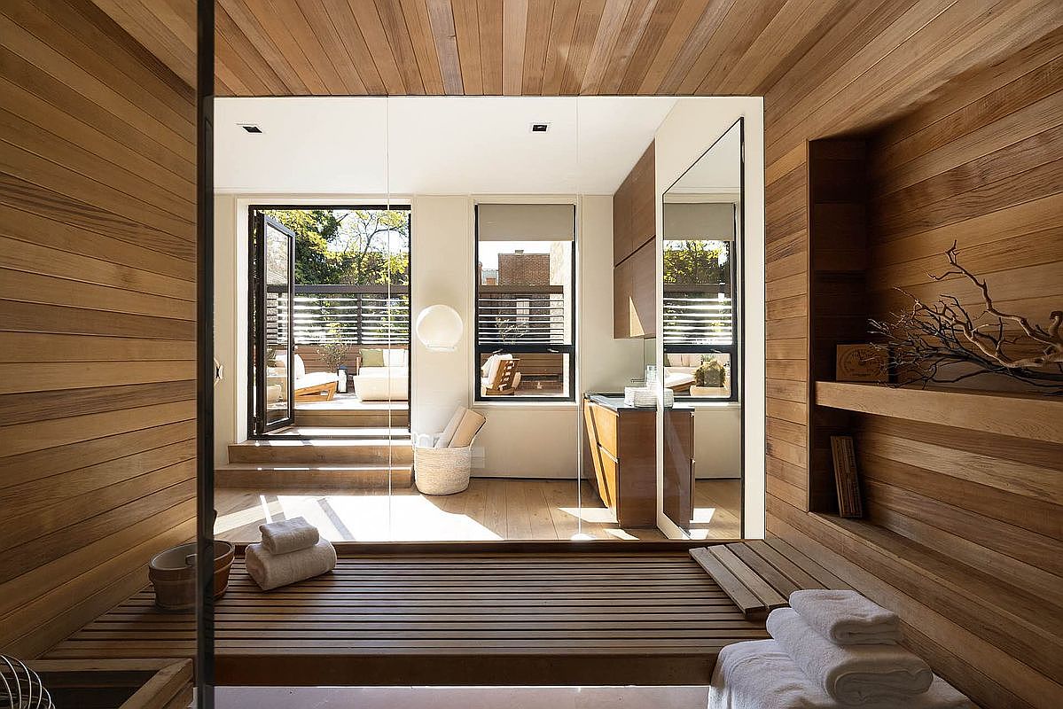 This is a luxurious primary bathroom featuring extensive wood paneling on the walls, ceiling, and floor, creating a warm and spa-like atmosphere. The bathroom opens to an outdoor patio with seating, accessible through a glass door. A modern vanity with a mirror and a built-in shelf add to the room's functionality and aesthetic appeal.