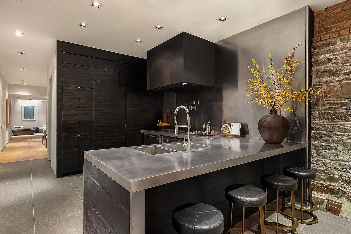 This is a modern kitchen with dark wood cabinetry and a stainless steel countertop. The kitchen features a large island with bar seating and a unique exposed brick wall. The overall impression is sleek and sophisticated, with a blend of modern and rustic elements.